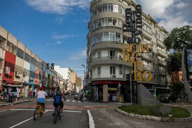 Ciclistas passam por uma rua movimentada do bairro Bom Retiro, em São Paulo, com destaque para o letreiro vertical de boas-vindas e edifícios comerciais e residenciais ao fundo sob céu azul.